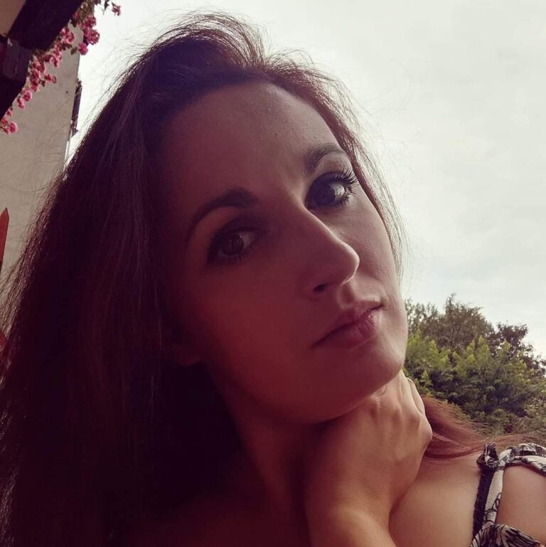 Zdjęcie pani Elżbiety Kowalskiej. Kobieta pozuje na zewnątrz w pochmurny dzień, opierając rękę na szyi. W tle widoczne są liście drzew i fragment budynku z wiszącymi różowymi kwiatami.
